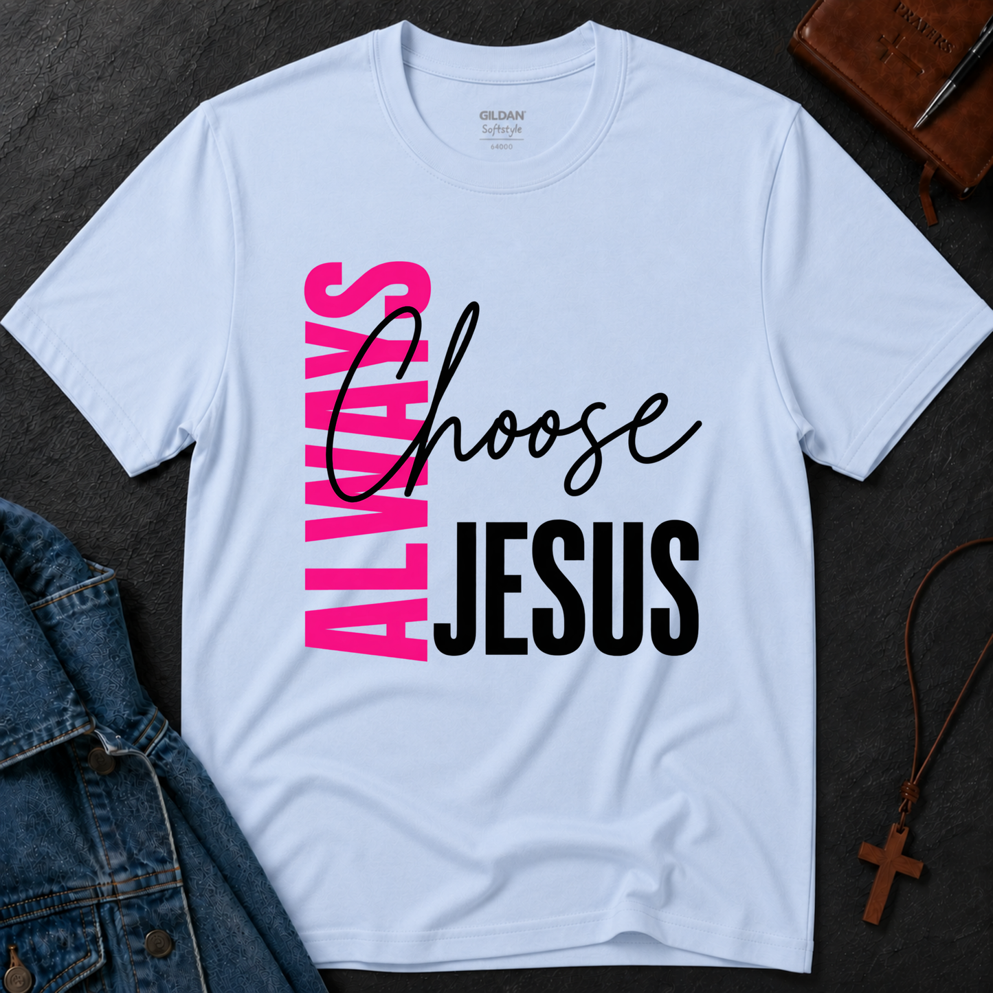 A light blue Gildan t-shirt laid flat on a dark textured surface, featuring a graphic print that reads 'ALWAYS Choose JESUS' in pink, black, and white. Styled flat-lay props include a denim jacket, a brown leather cross necklace, and a brown journal.