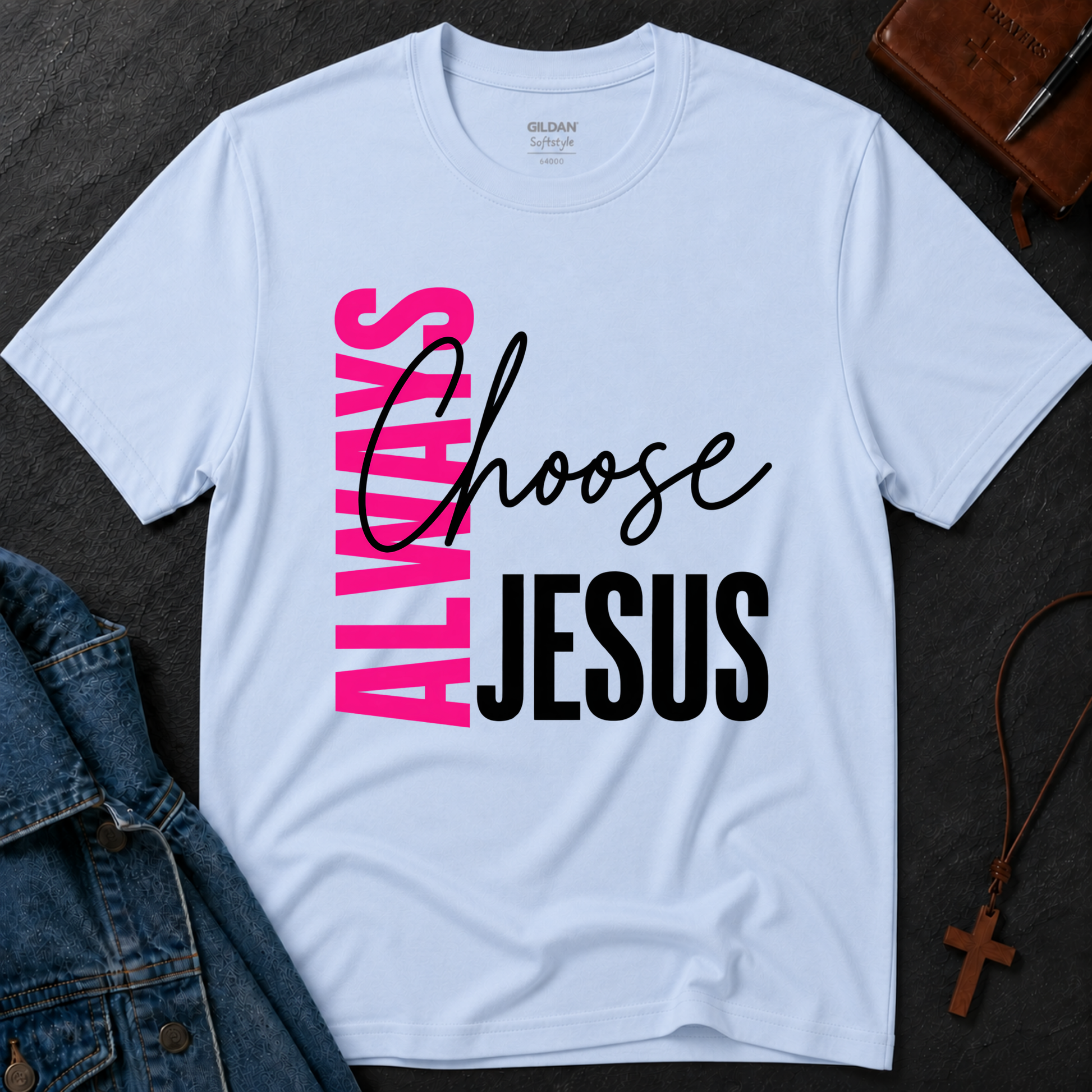 A light blue Gildan t-shirt laid flat on a dark textured surface, featuring a graphic print that reads 'ALWAYS Choose JESUS' in pink, black, and white. Styled flat-lay props include a denim jacket, a brown leather cross necklace, and a brown journal.