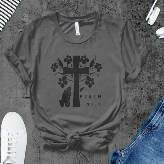 Gray t-shirt with black cross and floral design on a textured surface with jeans and sneakers.