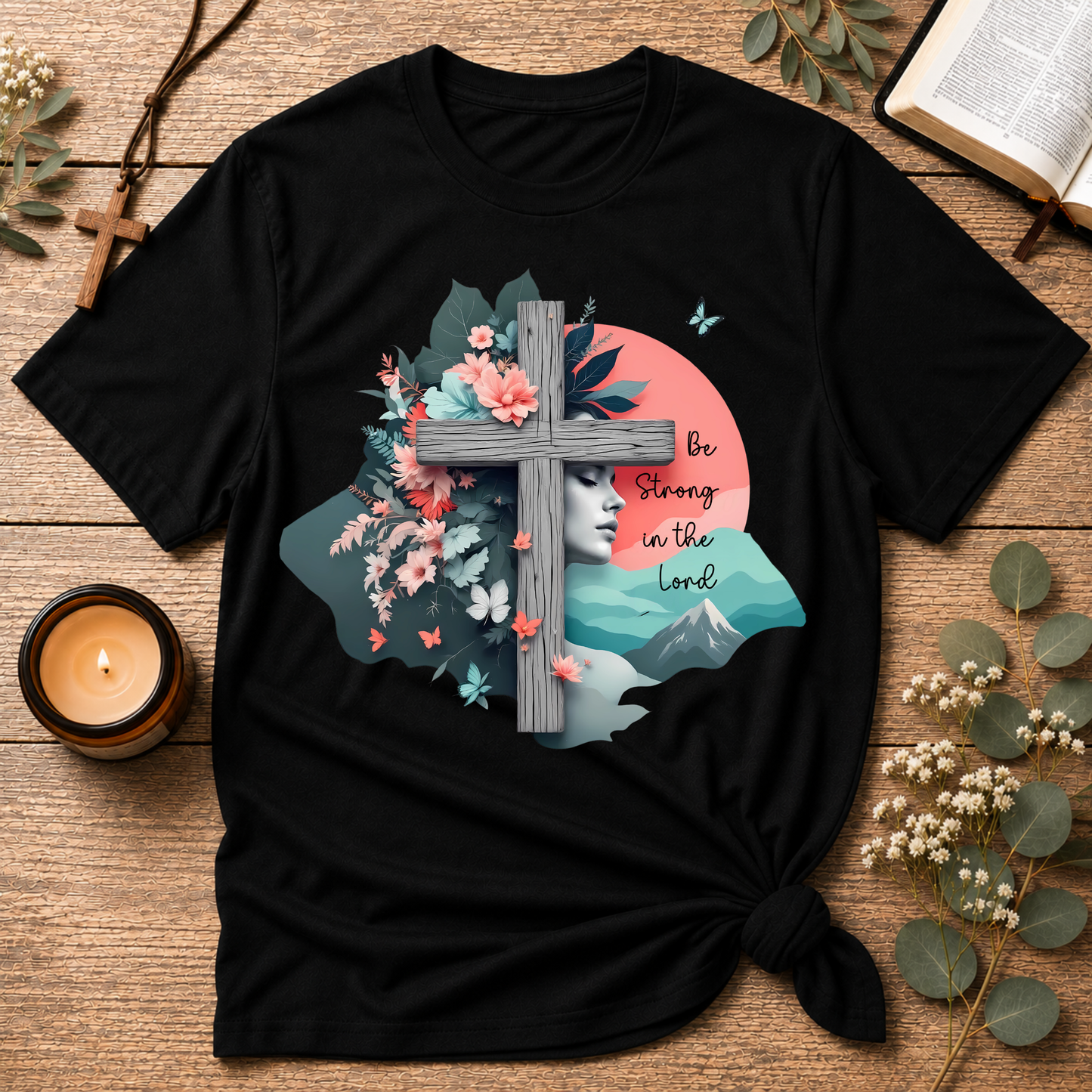 A black short-sleeve graphic T-shirt laid flat on a rustic wooden surface, styled with a candle, book pages, eucalyptus leaves, and a cross. The shirt features a large front print with a gray cross, pink flowers, teal leaves, a pale face, mountains, butterflies, and the text 'Be Strong in the Lord.