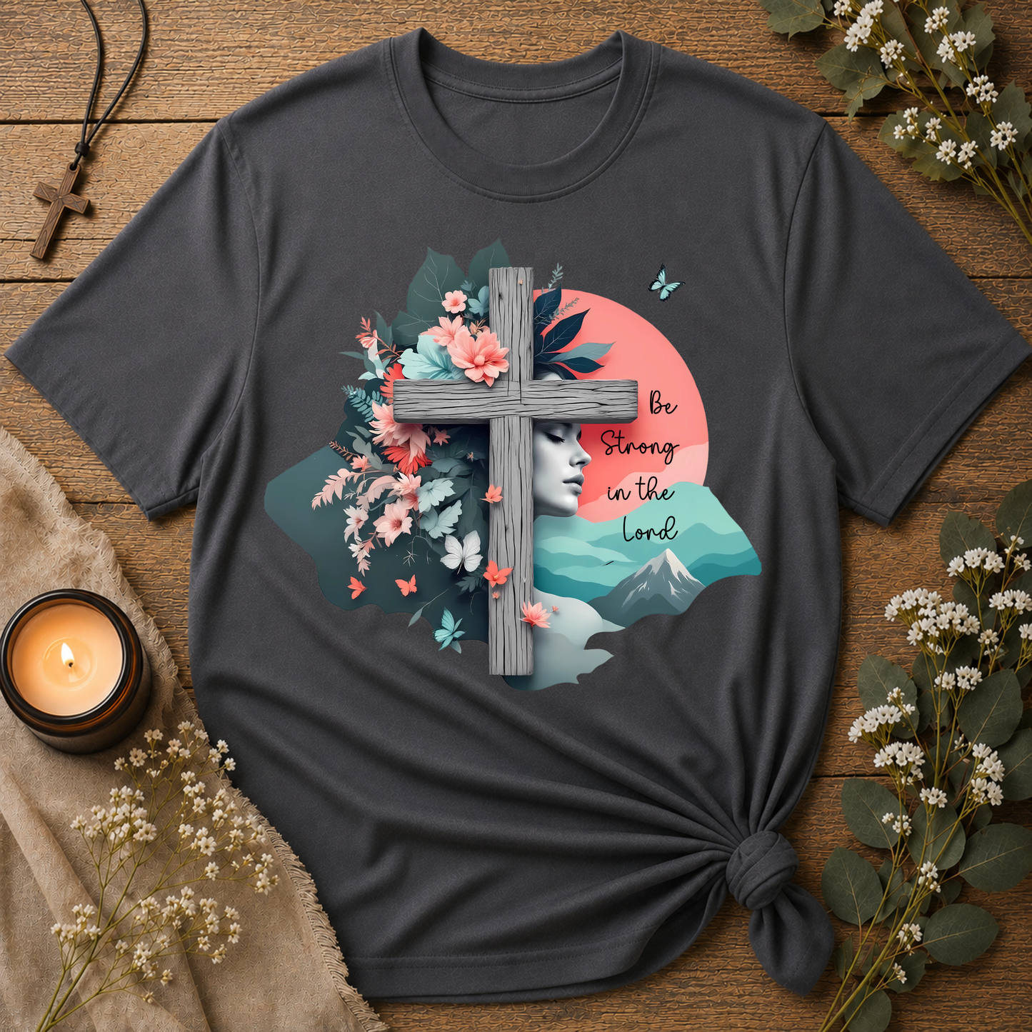 A dark charcoal gray graphic t-shirt styled in a flat-lay on a rustic wooden table. The t-shirt features a detailed graphic print with a wooden cross, a woman's profile, mountains, flowers, butterflies, and the text 'Be Strong in the Lord'.
