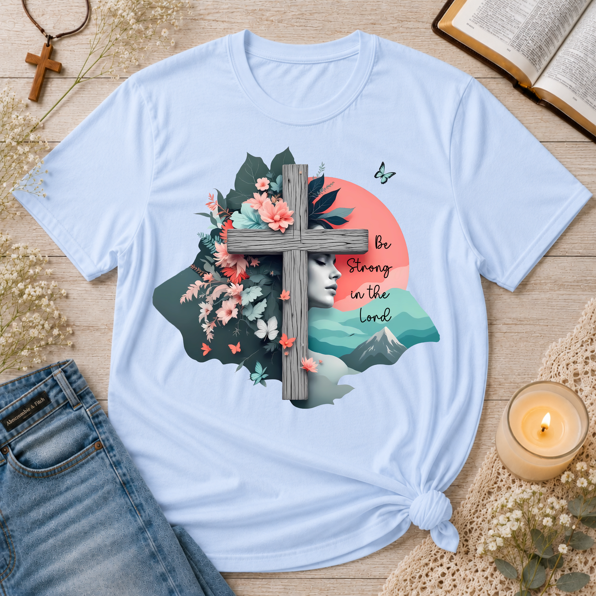 A light blue graphic t-shirt is displayed in a styled flat-lay arrangement on a wooden surface, surrounded by decorative props including an open book, a candle, jeans, and a cross. The t-shirt features a detailed graphic print with a wooden cross, pink flowers, a woman's face, mountains, and the text 'Be Strong in the Lord'.