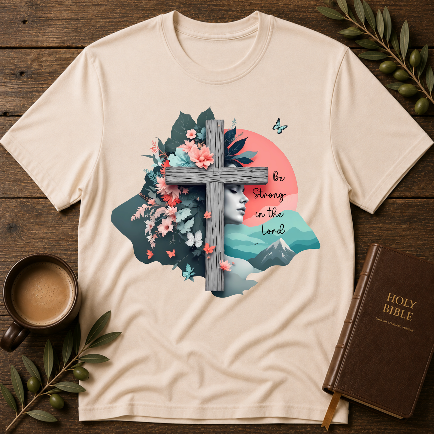 A cream-colored graphic t-shirt laid flat on a rustic wooden table, surrounded by a coffee cup, green leaves, and a brown leather-bound Holy Bible. The t-shirt features a detailed graphic of a woman's profile, a wooden cross, flowers, butterflies, mountains, and the text 'Be Strong in the Lord'.