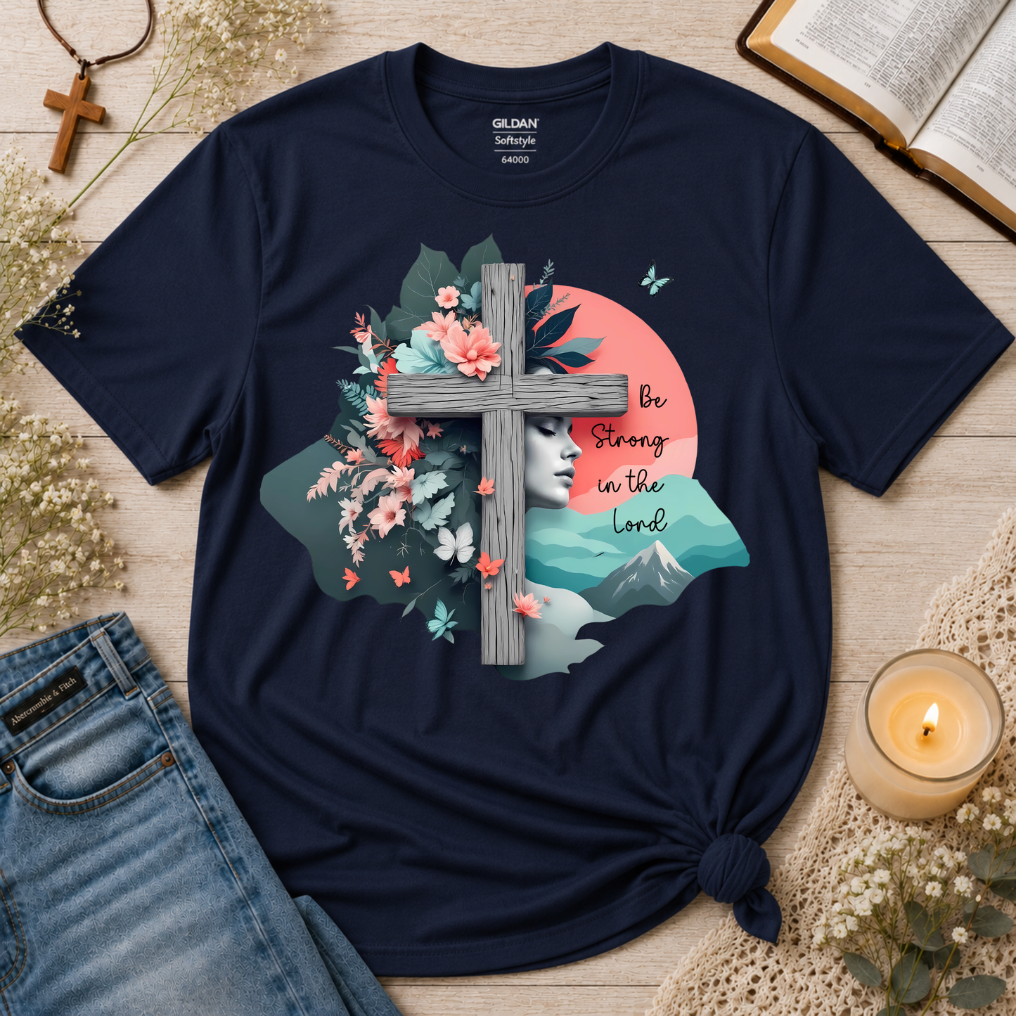A navy blue short-sleeve graphic T-shirt laid flat on a light wood surface with styled props including jeans, a candle, an open book, and dried flowers. The shirt has a large front print featuring a gray cross, pink flowers, teal leaves, a pale face, mountains, butterflies, and the text 'Be Strong in the Lord.