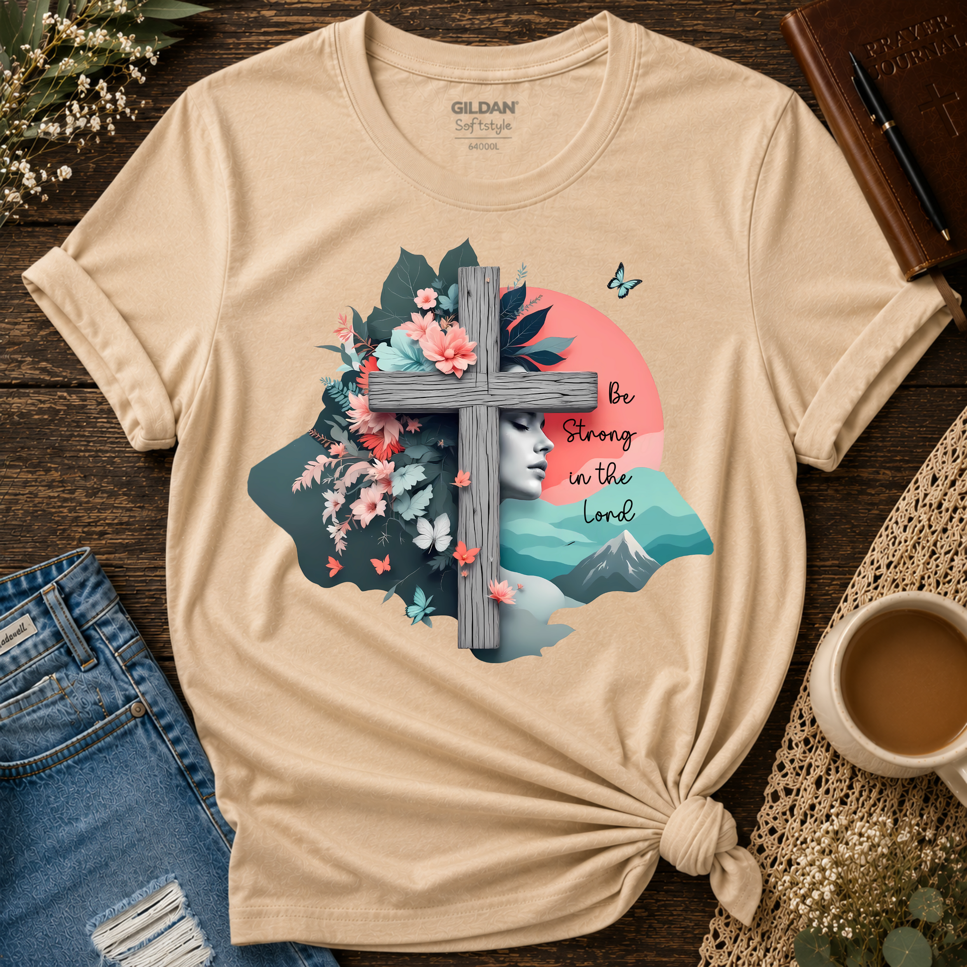 A beige Gildan Self-Style t-shirt with a graphic print featuring a wooden cross, flowers, a woman's face, mountains, and the text 'Be Strong in the Lord'. The shirt is styled in a flat-lay on a dark wood background with jeans, a coffee cup, and a book.