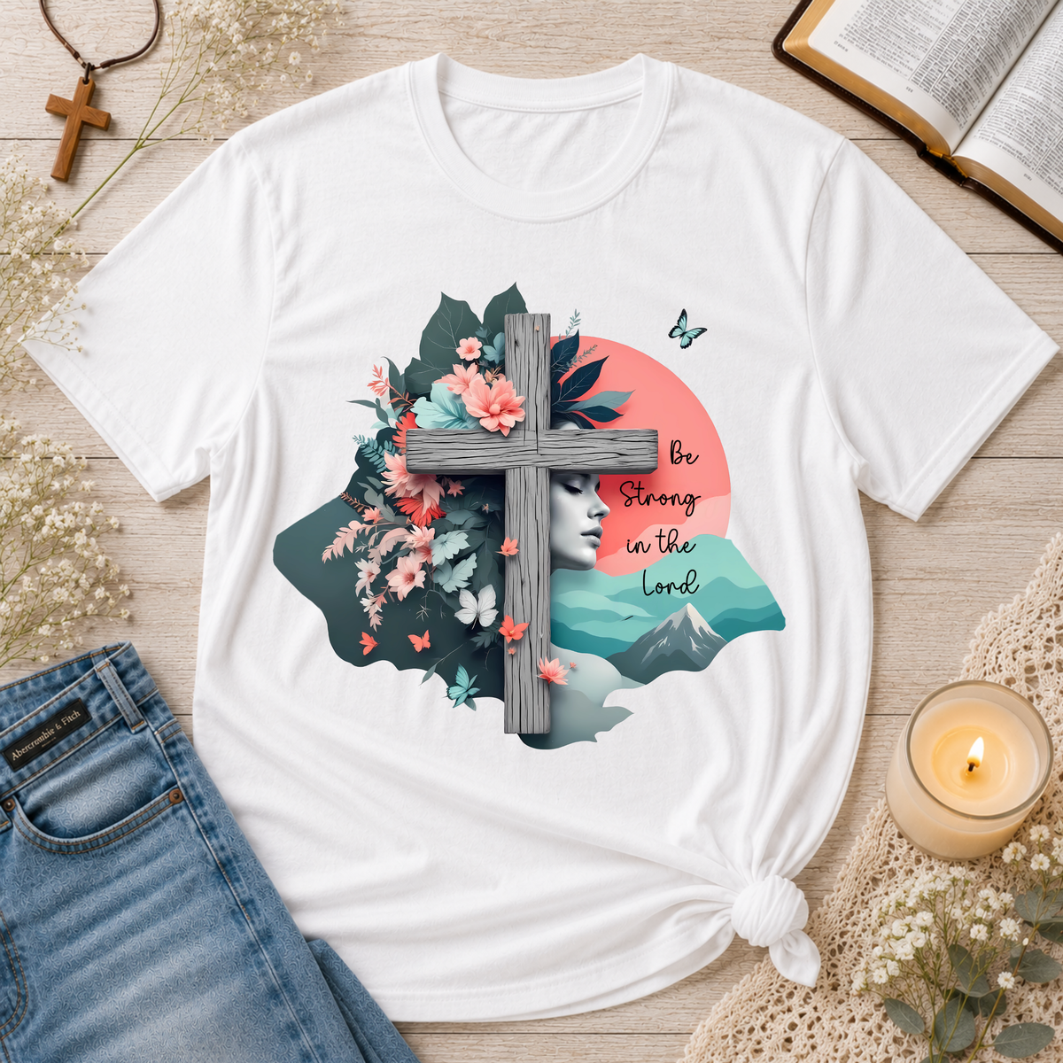 A white short-sleeve T-shirt laid flat on a light wood surface, styled with blue jeans, a candle, an open book, and small floral props. The shirt has a large front graphic featuring a wooden cross over a scenic landscape with flowers, butterflies, a woman's profile, and the text 'Be Strong in the Lord.