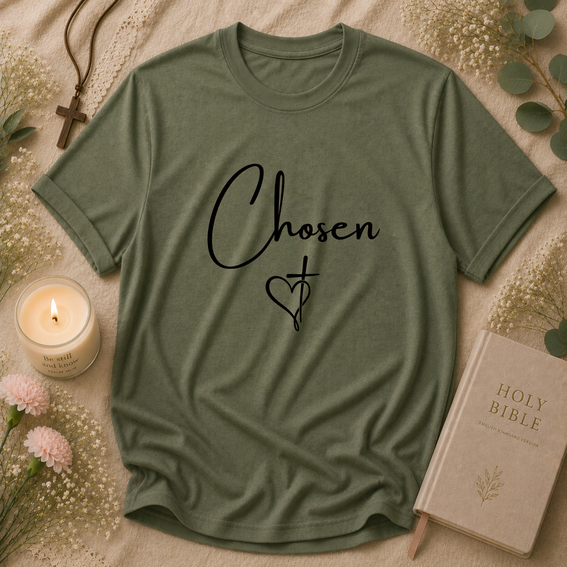 An military green short-sleeve t-shirt with the word 'Chosen' and a cross-heart graphic printed in black on the front. The t-shirt is styled in a flat-lay arrangement on a beige textured blanket, surrounded by a lit candle, a holy bible, flowers, and a necklace.