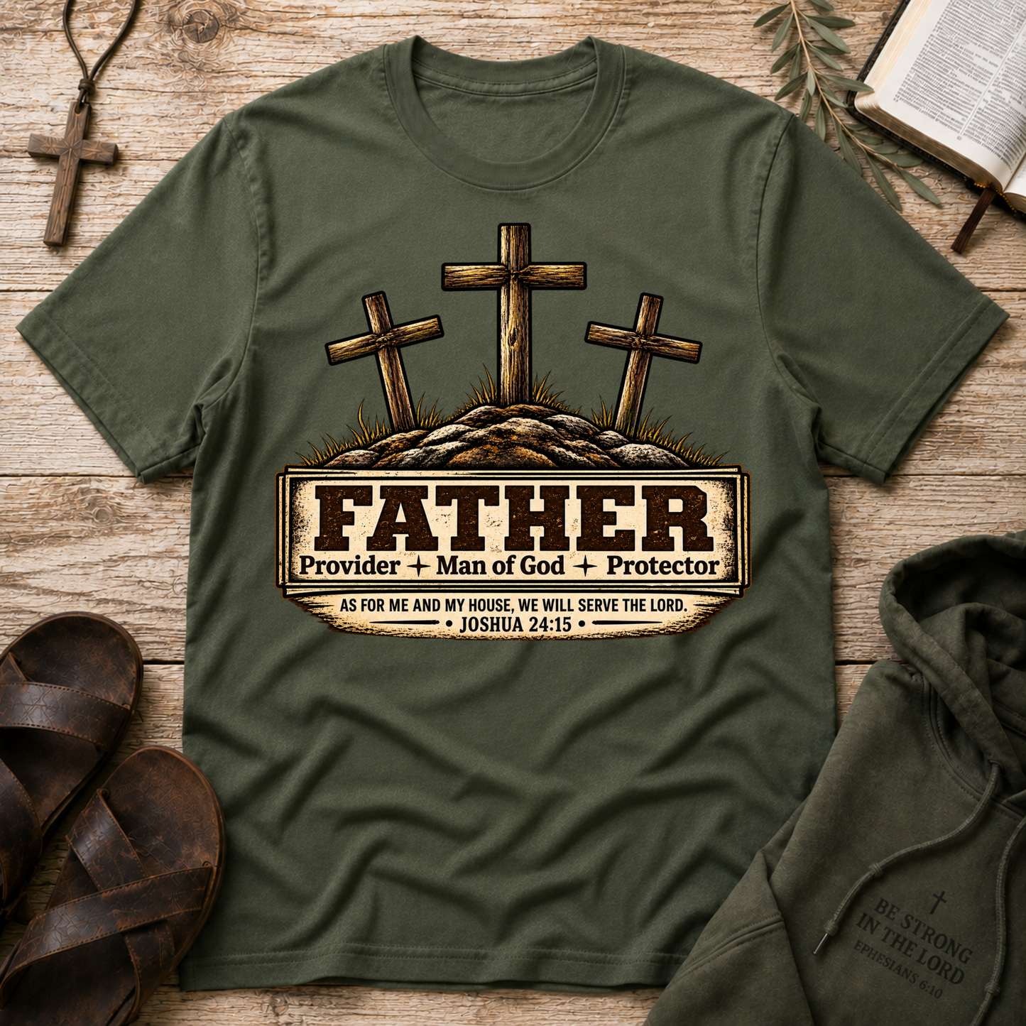 An military green graphic t-shirt laid flat on a rustic wooden background, styled with a brown cross necklace, sandals, a book, and a hoodie. The t-shirt features a graphic of three wooden crosses on a hill with a sign that reads 'FATHER Provider + Man of God + Protector' and a Bible verse from Joshua 24:15.