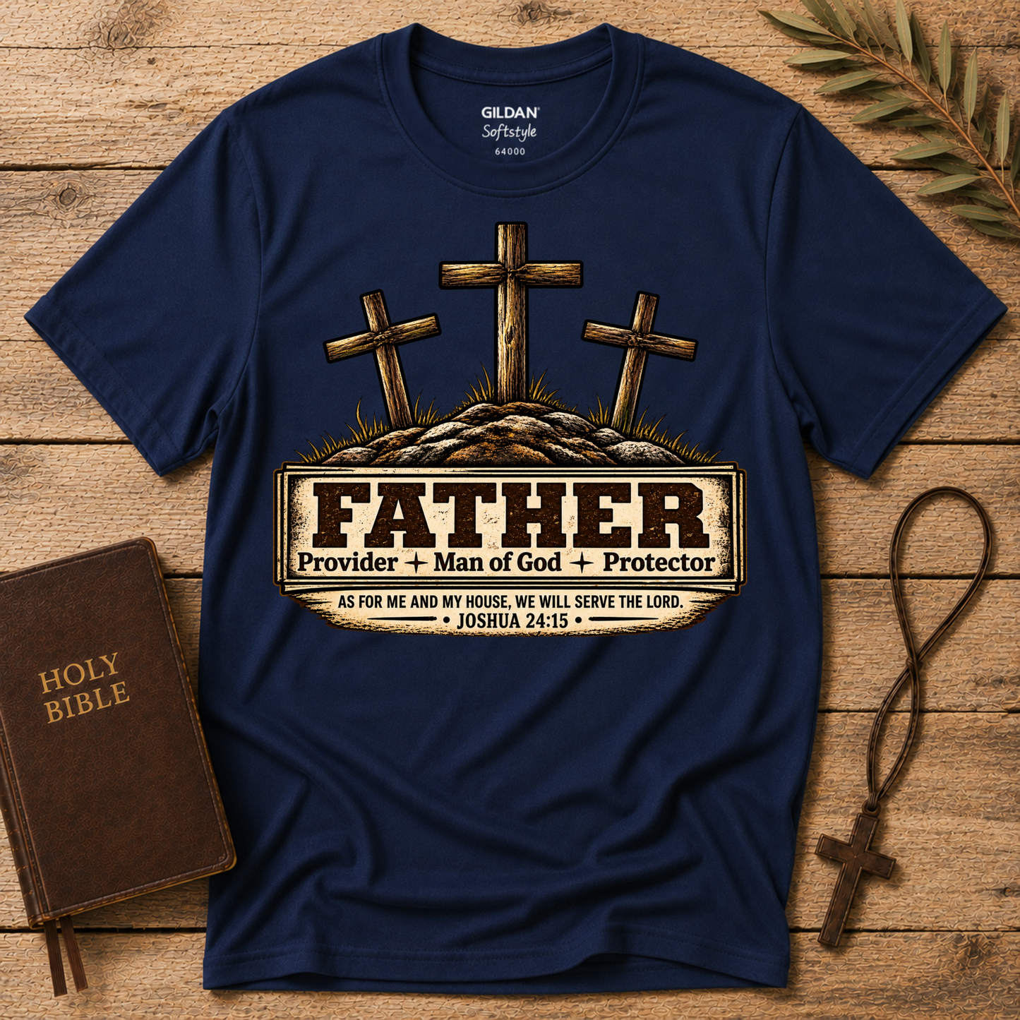 A navy blue Gildan Softstyle t-shirt with a graphic of three wooden crosses and a banner reading 'FATHER Provider + Man of God + Protector' with a Bible verse, styled on a wooden background with a Holy Bible, a rosary, and a palm leaf.