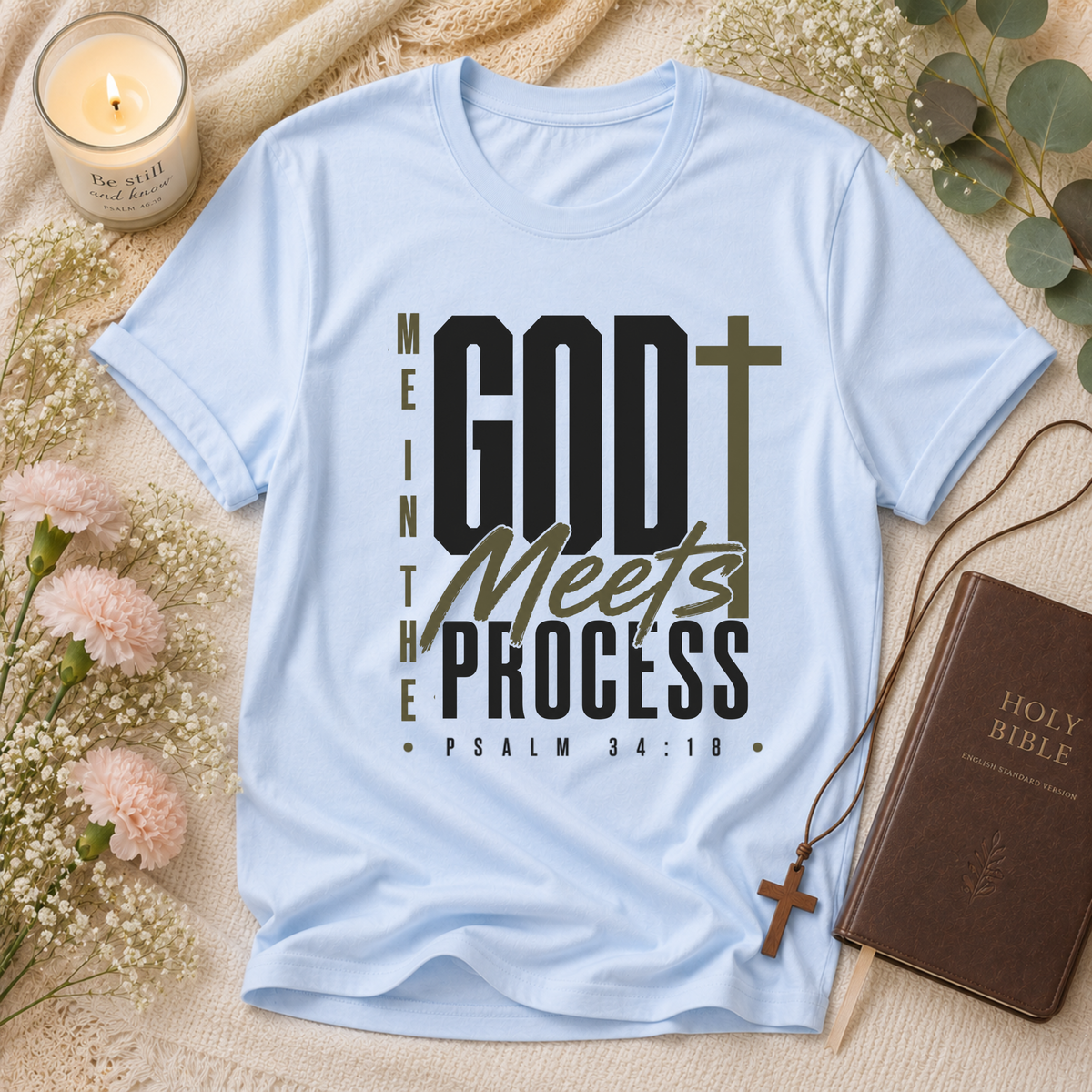 A light blue graphic t-shirt laid flat on a beige fabric background, surrounded by decorative props including a lit candle, flowers, a Holy Bible, and a cross necklace. The shirt features the text 'MEANWHILE GOD Meets PROCESS PSALM 34:18' in black and olive green lettering.