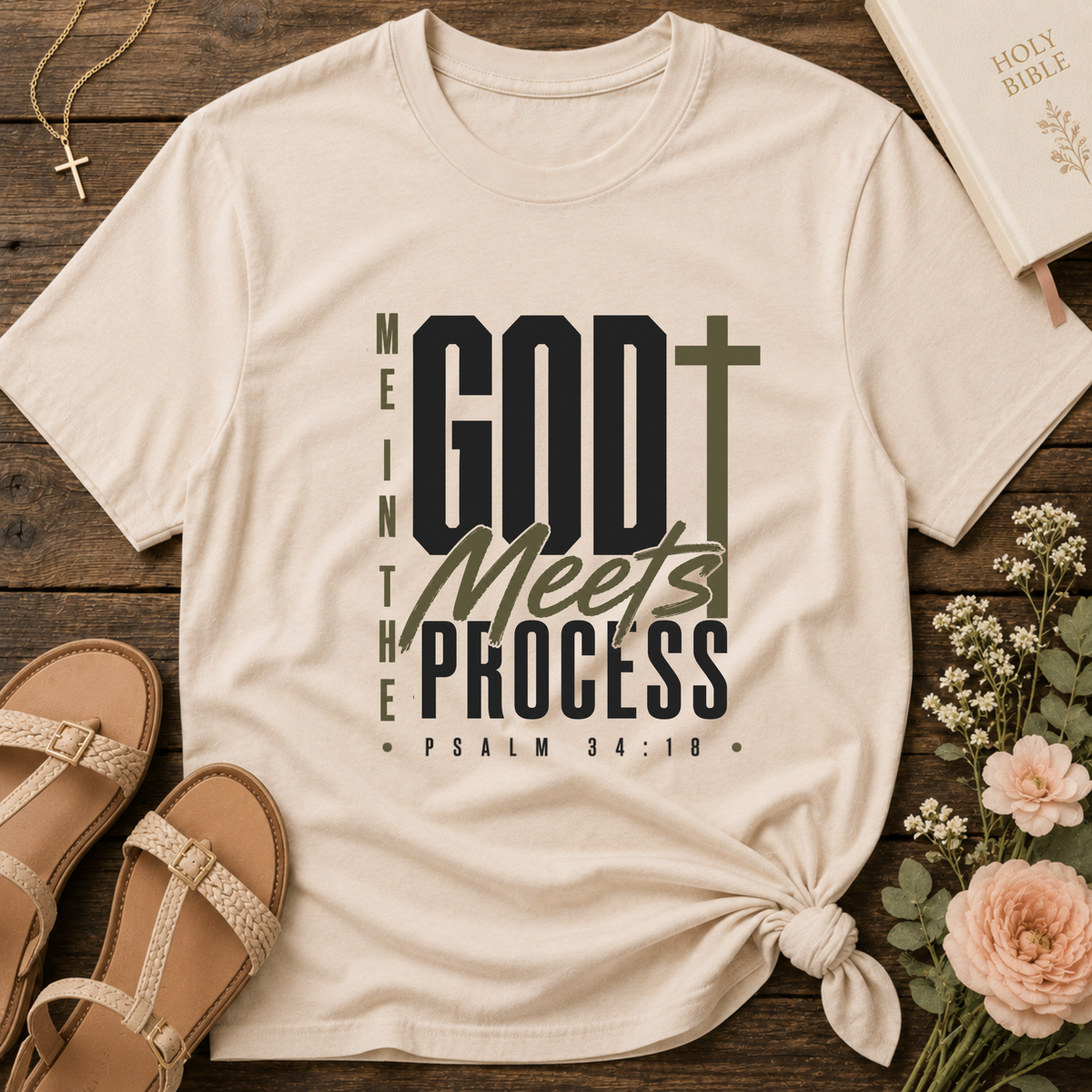A cream-colored graphic t-shirt with a knotted hem, displayed in a flat-lay arrangement on a rustic wooden background alongside sandals, a Bible, flowers, and a necklace. The t-shirt features the text 'GOD Meets THE PROCESS' with a cross graphic.