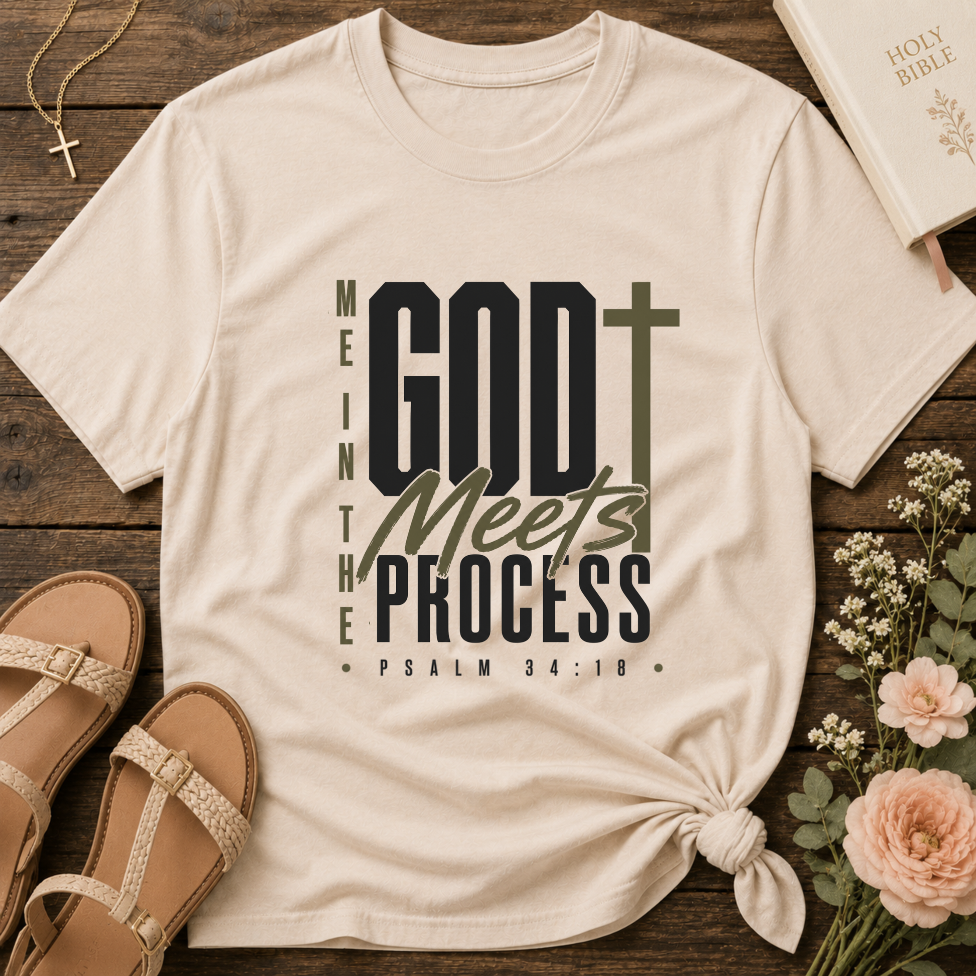 A cream-colored graphic t-shirt with a knotted hem, displayed in a flat-lay arrangement on a rustic wooden background alongside sandals, a Bible, flowers, and a necklace. The t-shirt features the text 'GOD Meets THE PROCESS' with a cross graphic.