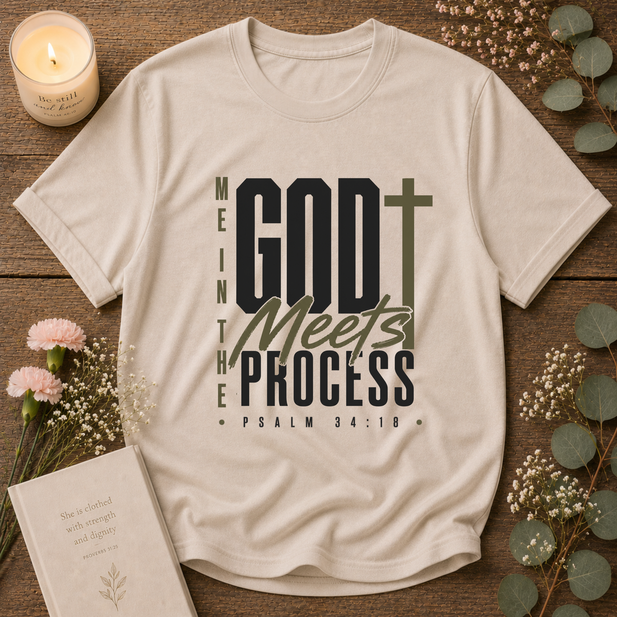 A beige graphic T-shirt laid flat on a rustic wooden surface with decorative flowers, a candle, and a book. The shirt has a large front print reading “ME IN THE GOD T Meets PROCESS PSALM 34:18” in black and olive green lettering with a cross symbol.