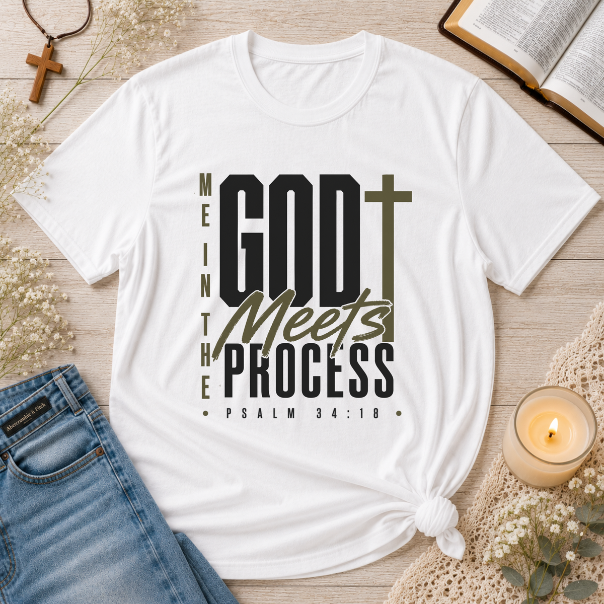 A white graphic t-shirt with the text 'ME IN THE GOD Meets THE PROCESS PSALM 34:18' in black and olive green, styled in a flat-lay arrangement with blue jeans, a lit candle, an open book, and a cross on a wooden surface.