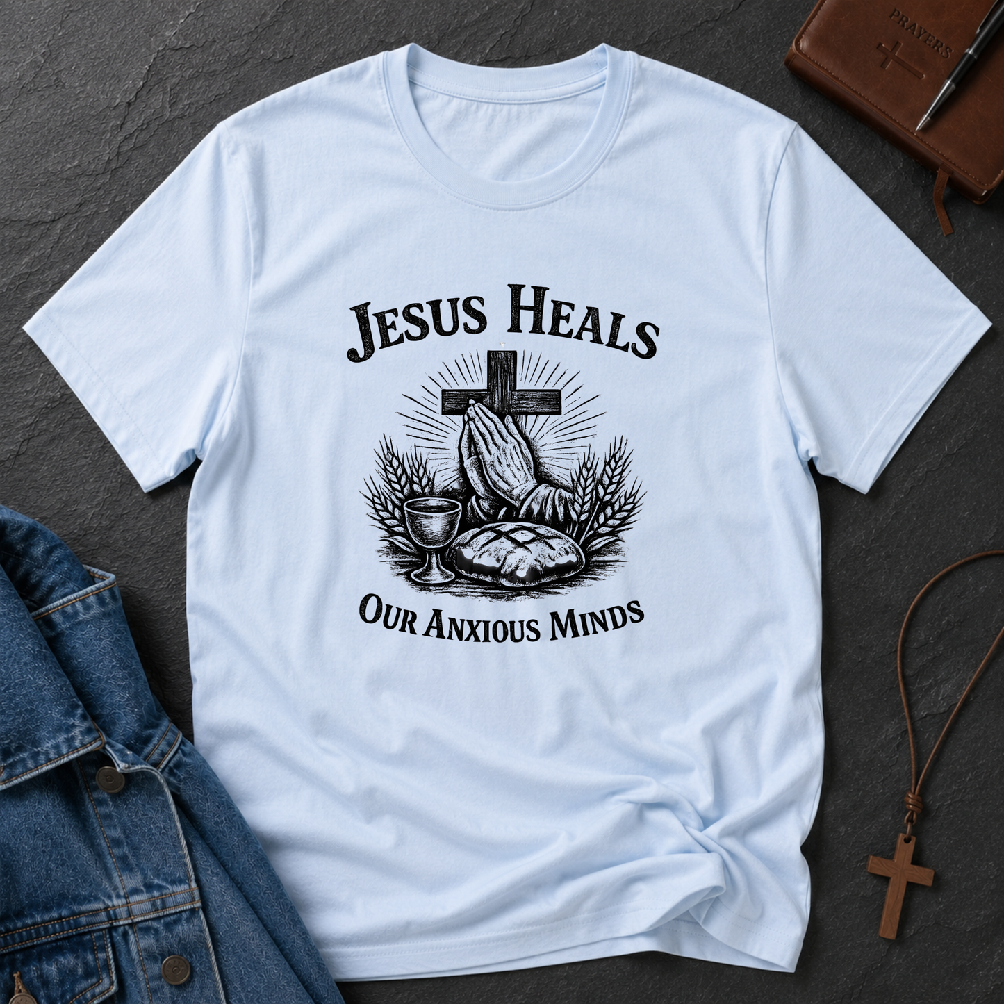 A light blue short-sleeve graphic T-shirt laid flat on a dark textured surface, with a black printed illustration of praying hands, a cross, a chalice, and wheat, plus the text 'JESUS HEALS OUR ANXIOUS MINDS.' A denim jacket corner, a brown leather journal, and a wooden cross necklace are placed around it as styling props.
