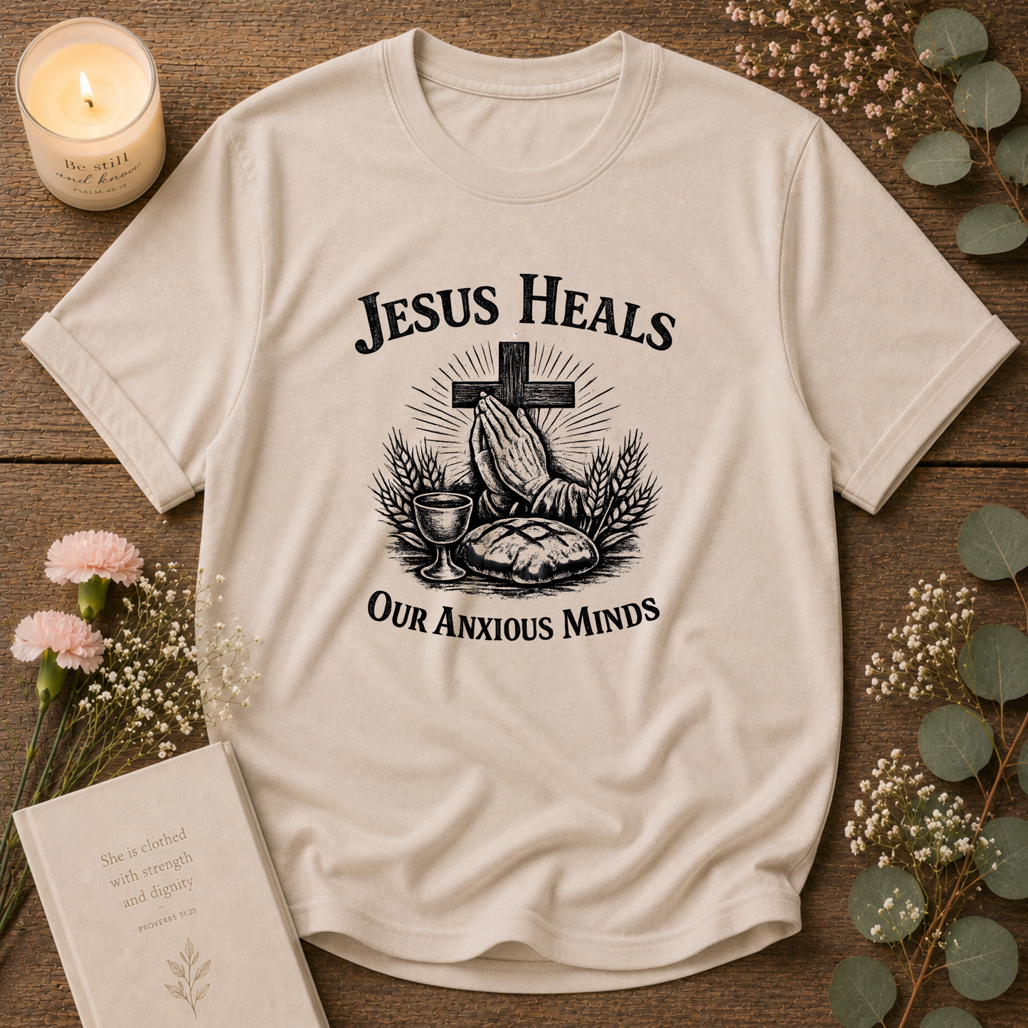 A cream-colored short-sleeve t-shirt with a black graphic print reading 'JESUS HEALS OUR ANXIOUS MINDS' around a cross, prayer hands, bread, and wine cup. The shirt is displayed in a flat-lay arrangement on a rustic wooden background, surrounded by a lit candle, a book, and small flowers.