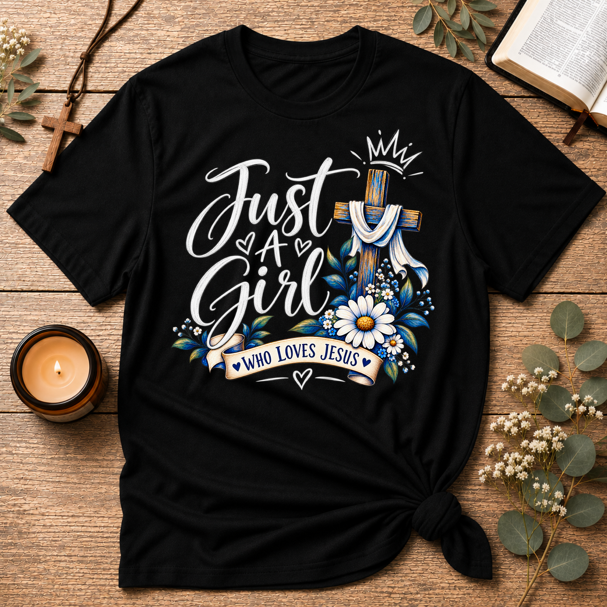 A black short-sleeve graphic T-shirt laid flat on a rustic wooden surface with decorative props including a candle, book pages, eucalyptus leaves, and a cross. The shirt features large white script text reading 'Just A Girl' with blue and white floral artwork, a blue-and-white cross, and a banner that says 'WHO LOVES JESUS'.