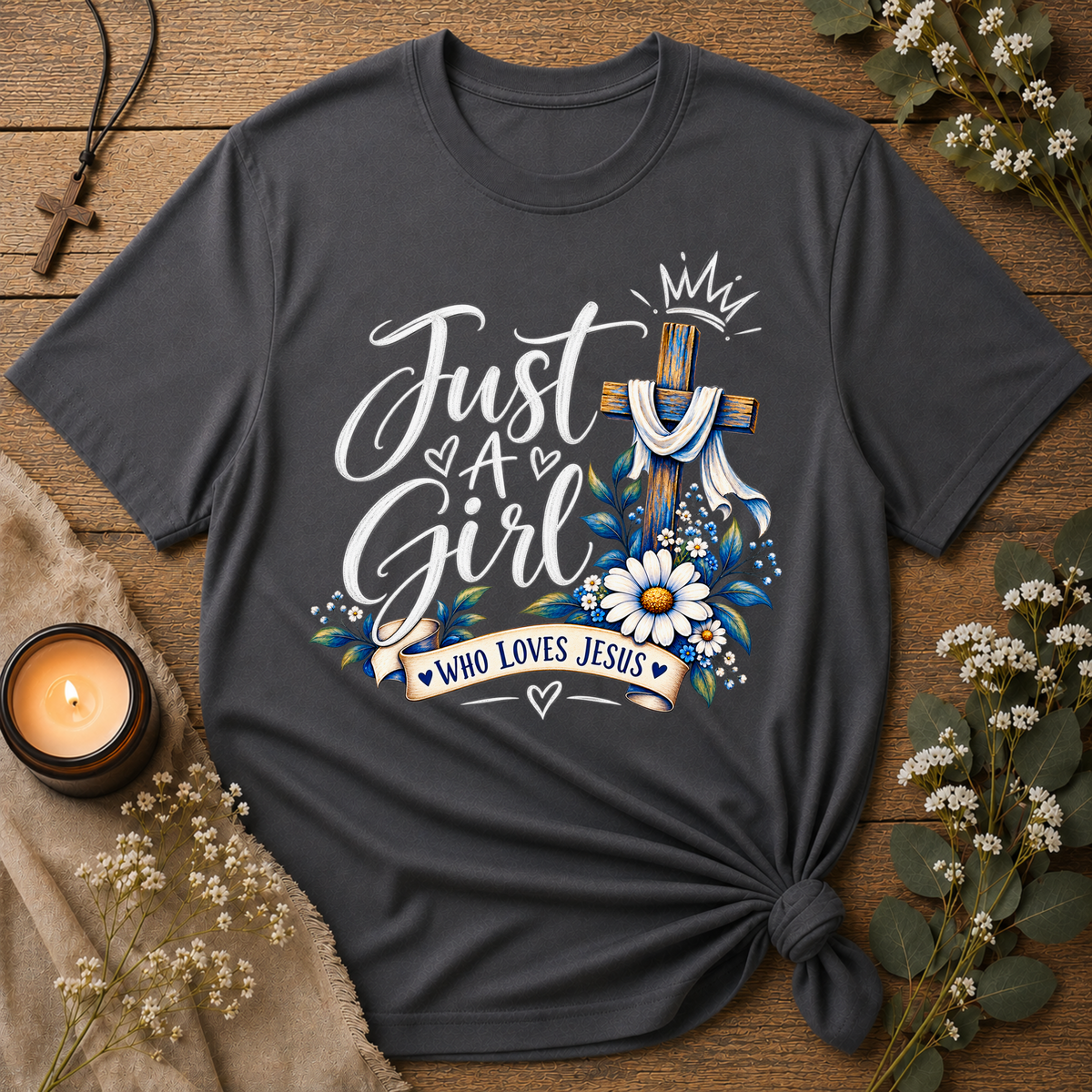 A dark charcoal gray graphic t-shirt styled in a flat-lay on a rustic wooden background, featuring a 'Just A Girl Who Loves Jesus' design with a cross, flowers, and hearts. The shirt is styled with a knotted hem and surrounded by decorative props including a candle, a cross necklace, and dried flowers.
