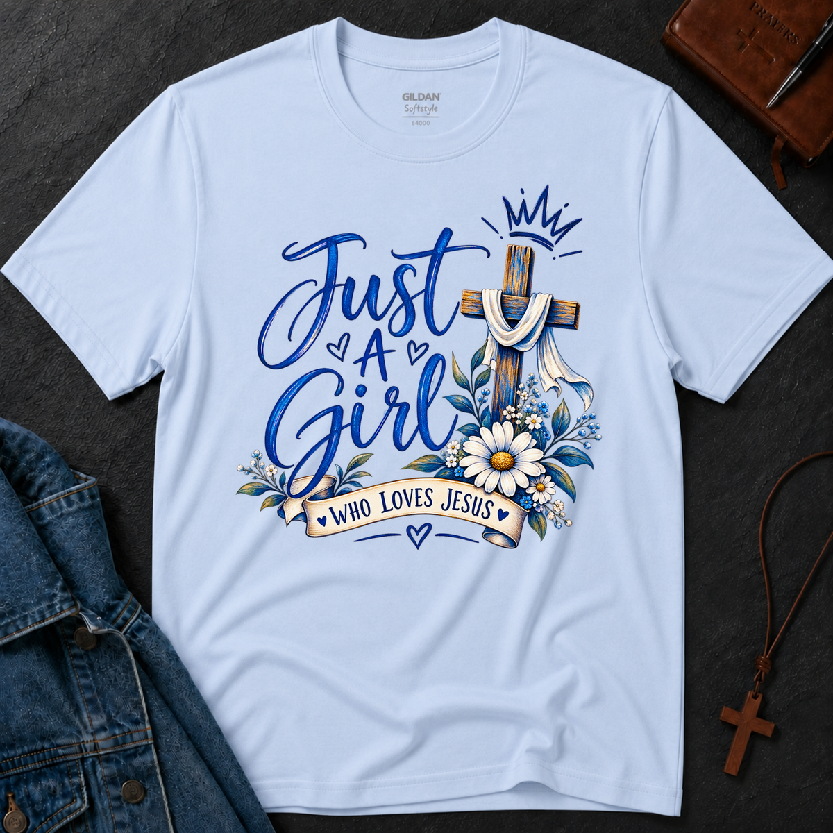 A light blue Gildan t-shirt laid flat on a dark textured surface, featuring a graphic design with the words 'Just A Girl Who Loves Jesus' in blue script, a wooden cross draped with a white cloth, white daisies, blue leaves, and a small crown. Styled flat-lay props include a denim jacket, a brown leather journal, and a wooden cross necklace.