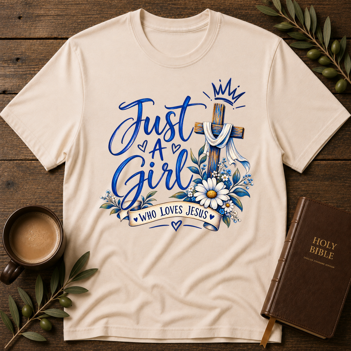A natural short-sleeve graphic T-shirt laid flat on a light wood surface, styled with jeans, a candle, an open book, and small floral decor. The shirt has a large front print reading “Just A Girl Who Loves Jesus” with a cross, crown, flowers, and hearts.