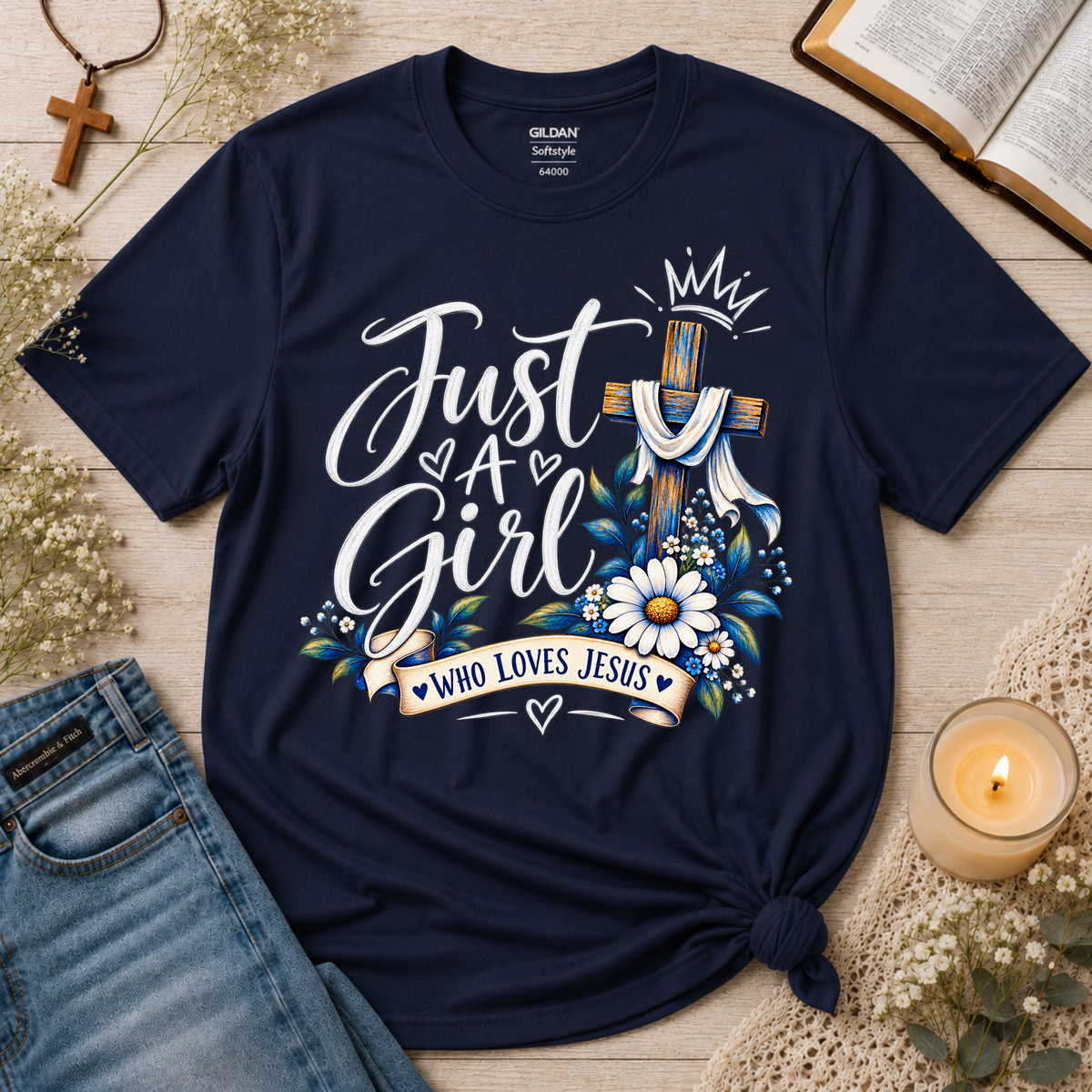 A navy blue short-sleeve graphic T-shirt laid flat on a light wood surface, styled with jeans, a candle, an open book, and small floral decor. The shirt has a large front print reading “Just A Girl Who Loves Jesus” with a cross, crown, flowers, and hearts.