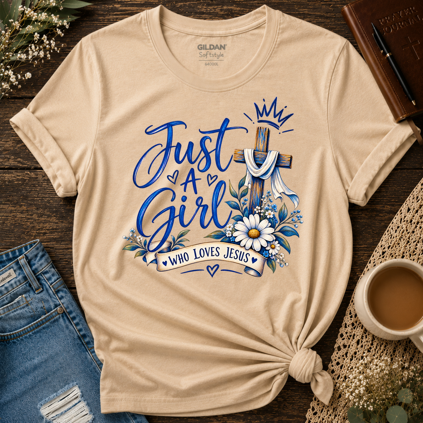 A beige Gildan Self-Style t-shirt with a graphic print reading 'Just A Girl Who Loves Jesus' in blue script, decorated with a wooden cross, white daisies, and a crown. The shirt is styled in a flat-lay on a dark wood background with jeans, a cup of coffee, and a book.