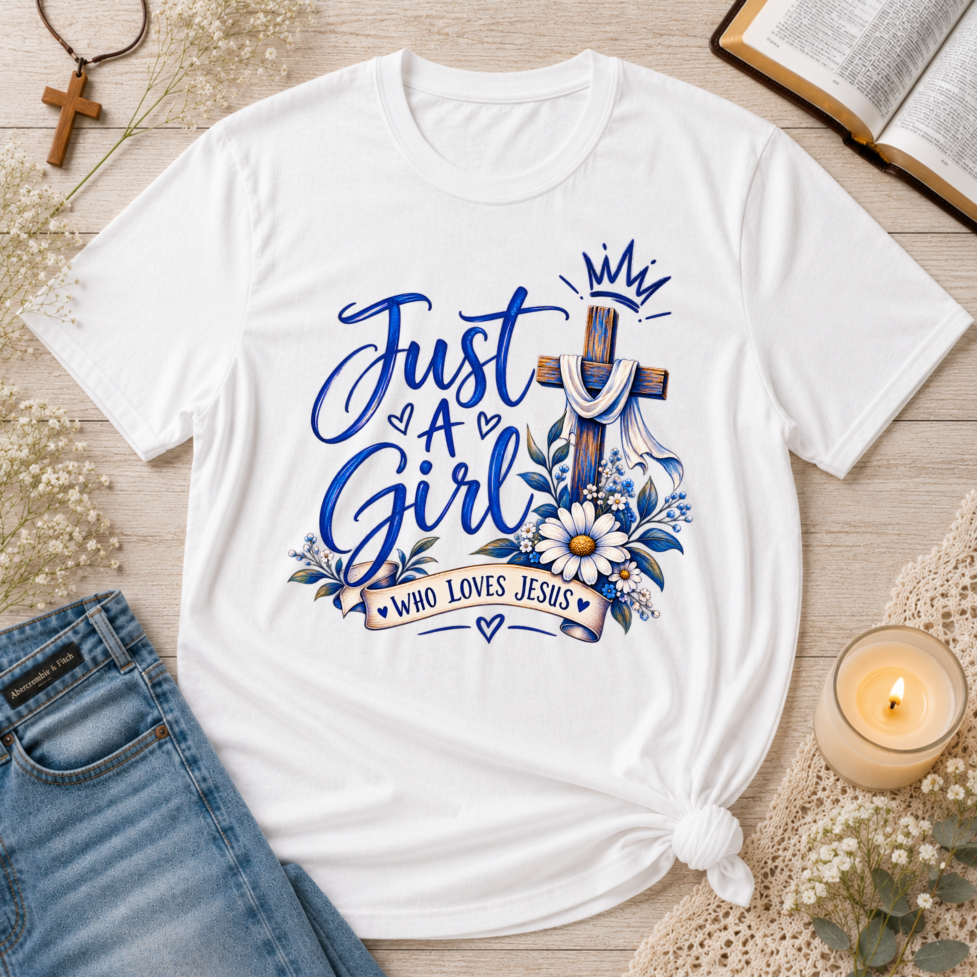 A white graphic T-shirt laid flat on a light wood surface with styled props including blue jeans, a candle, an open book, a cross necklace, and dried flowers. The shirt graphic reads “Just A Girl Who Loves Jesus” with a blue cross, crown, flowers, and hearts.