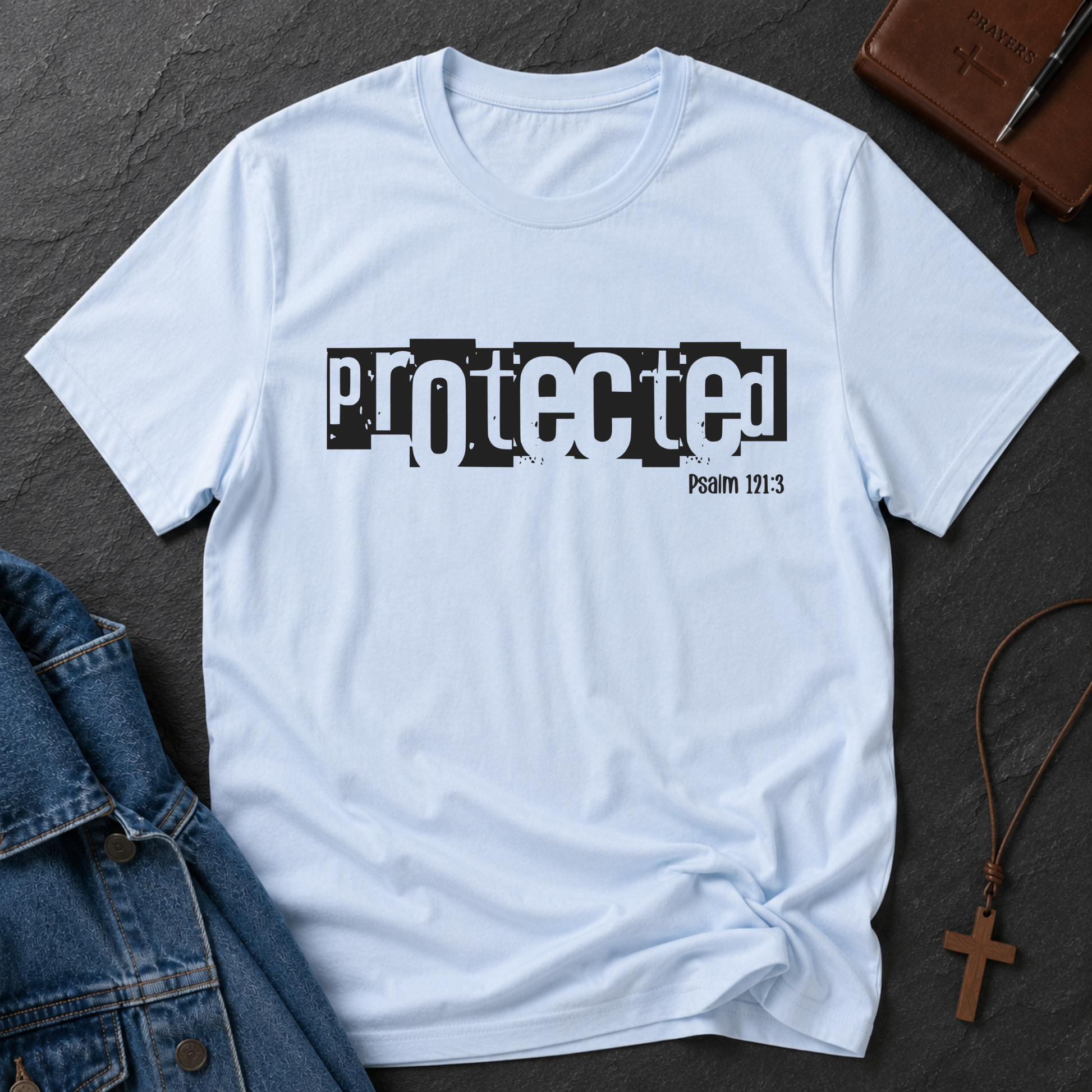 A light blue crewneck t-shirt with a black graphic print reading 'protected Psalm 171:3' in a distressed stencil-style font. The shirt is styled in a flat-lay arrangement on a dark gray slate background, surrounded by a blue denim jacket, a brown leather cross necklace, and a brown leather journal.