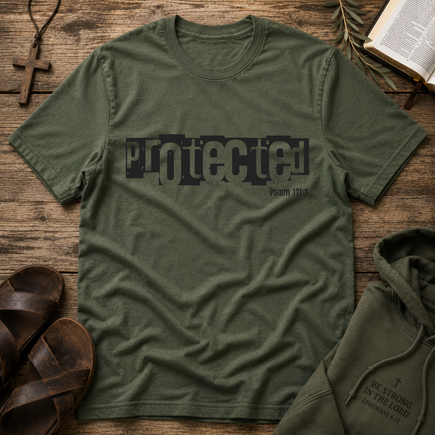 An military green crewneck t-shirt with a distressed black graphic reading 'protected' and 'Psalm 176:1' underneath, styled in a flat lay on a rustic wooden background with a hoodie, sandals, a cross, and an open book.