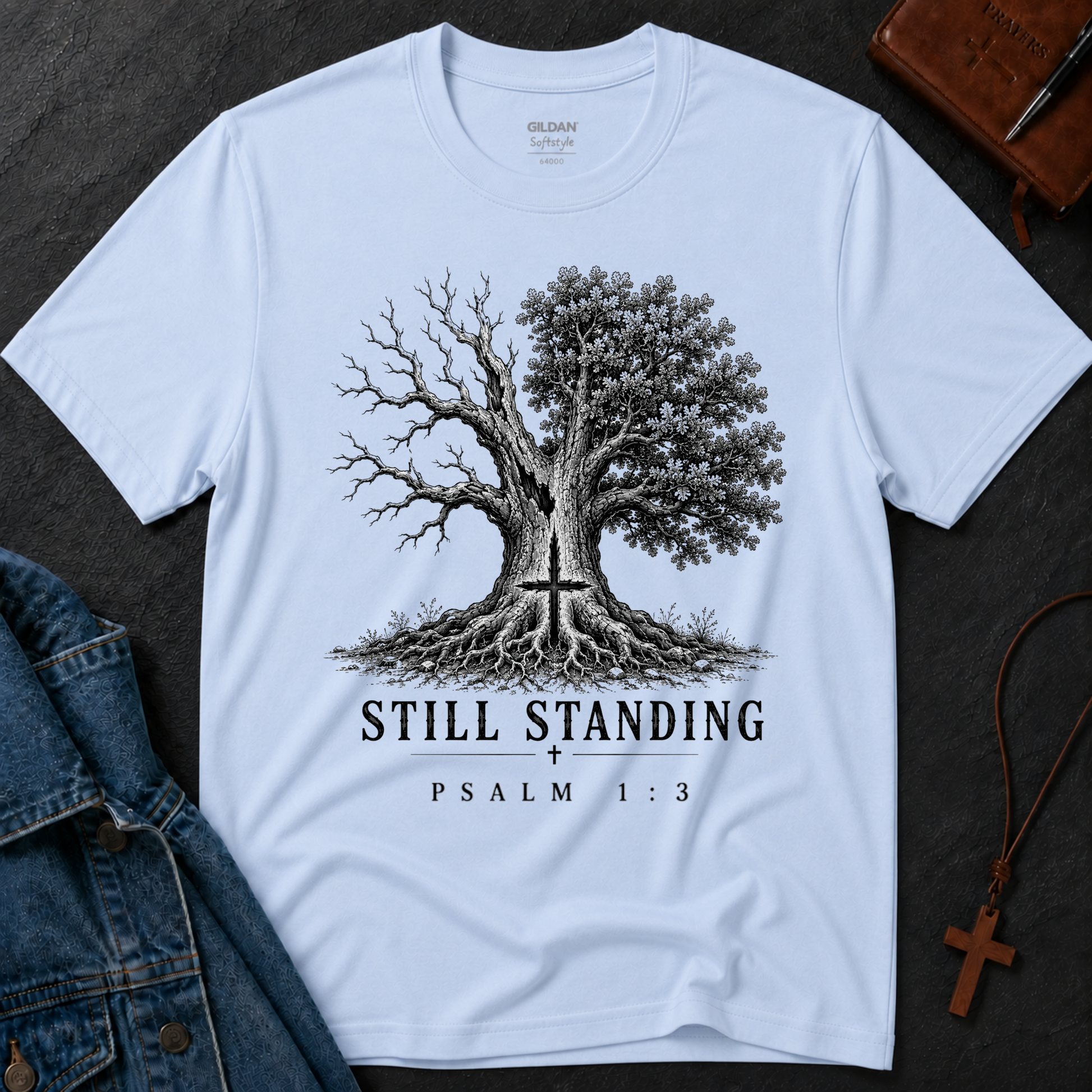 A light blue Gildan t-shirt laid flat on a dark textured surface, featuring a black and white graphic of a tree with one bare branch and one leafy canopy, both overlaid with a cross, with the text 'STILL STANDING' and 'PSALM 1:3' below. A denim jacket, a brown leather Bible, and a wooden cross necklace are styled around the shirt.